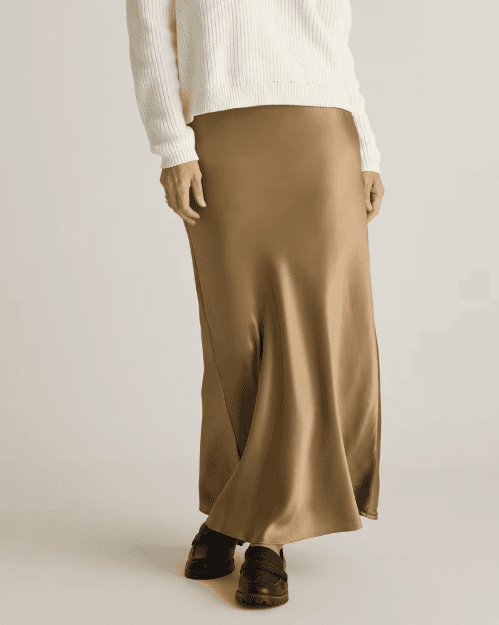 A person wearing a cream-colored ribbed sweater, a long gold satin skirt, and black loafers stands against a neutral background. The persons head is not visible in the image, by McGee & Co.